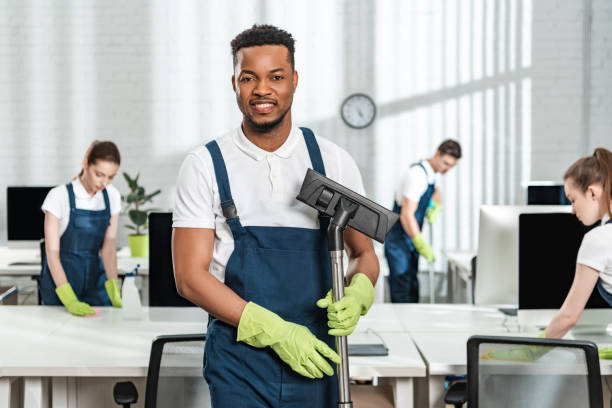 smiling african american cleaner holding vacuum cleaner brush near team of multicultural colleagues